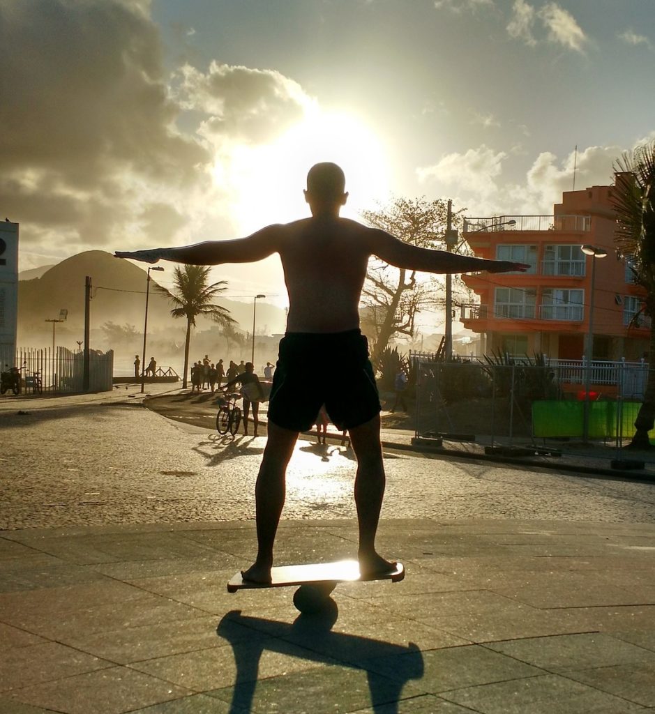 Ein Mann steht auf einem Balance Board und wird von hinten durch die Sonne angestrahlt, sodass man nur die Umrisse erkennen kann. Im Hintergrund sind zudem noch eine Straße, Menschen und Häuser zu erkennen. 