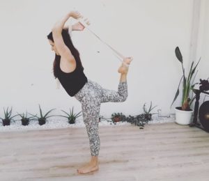 Eine Frau steht auf einem Bein und dehnt ihr anderes Bein mit der Hilfe eines Yoga Gurtes hinter dem Rücken. Im Hintergrund sind Pflanzen an der Wand aufgereiht.